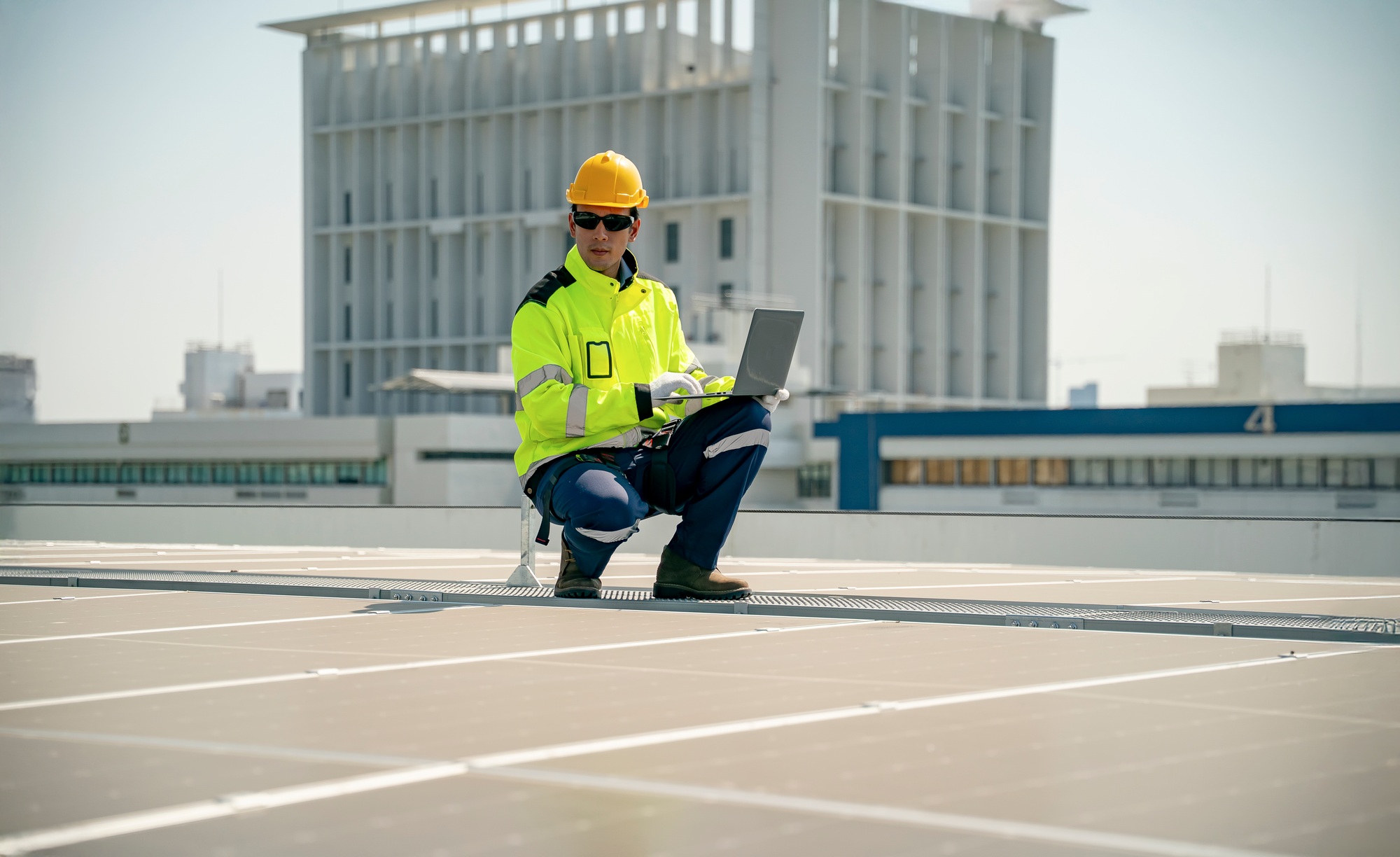 Worker inspects solar panels on rooftop while using a laptop in a city environment