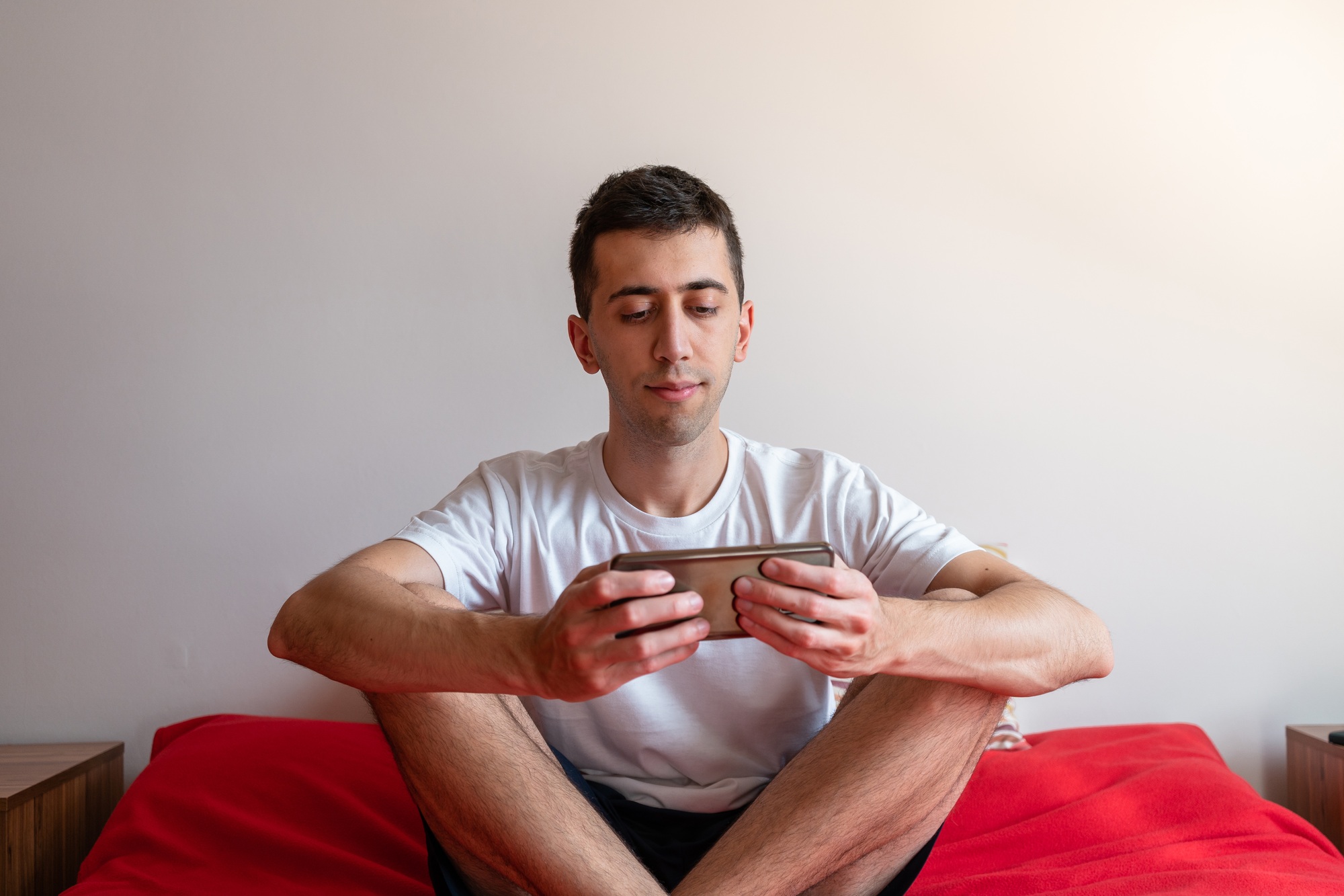 Young male playing with his smartphone while sitting on the bed.